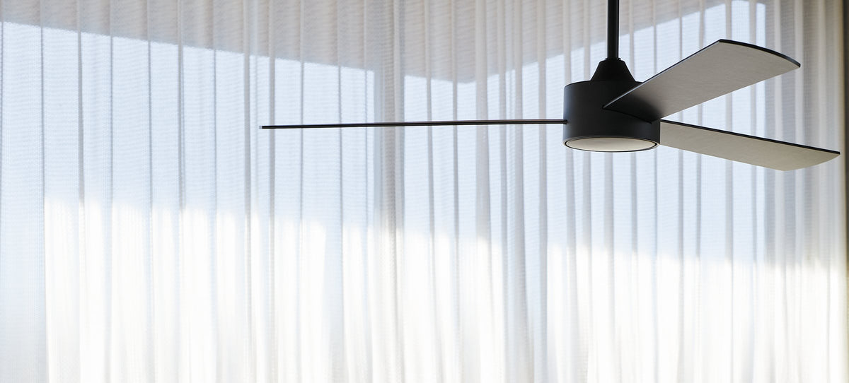 Black ceiling fan with pale timber blades in front of floor-length sheer white curtains