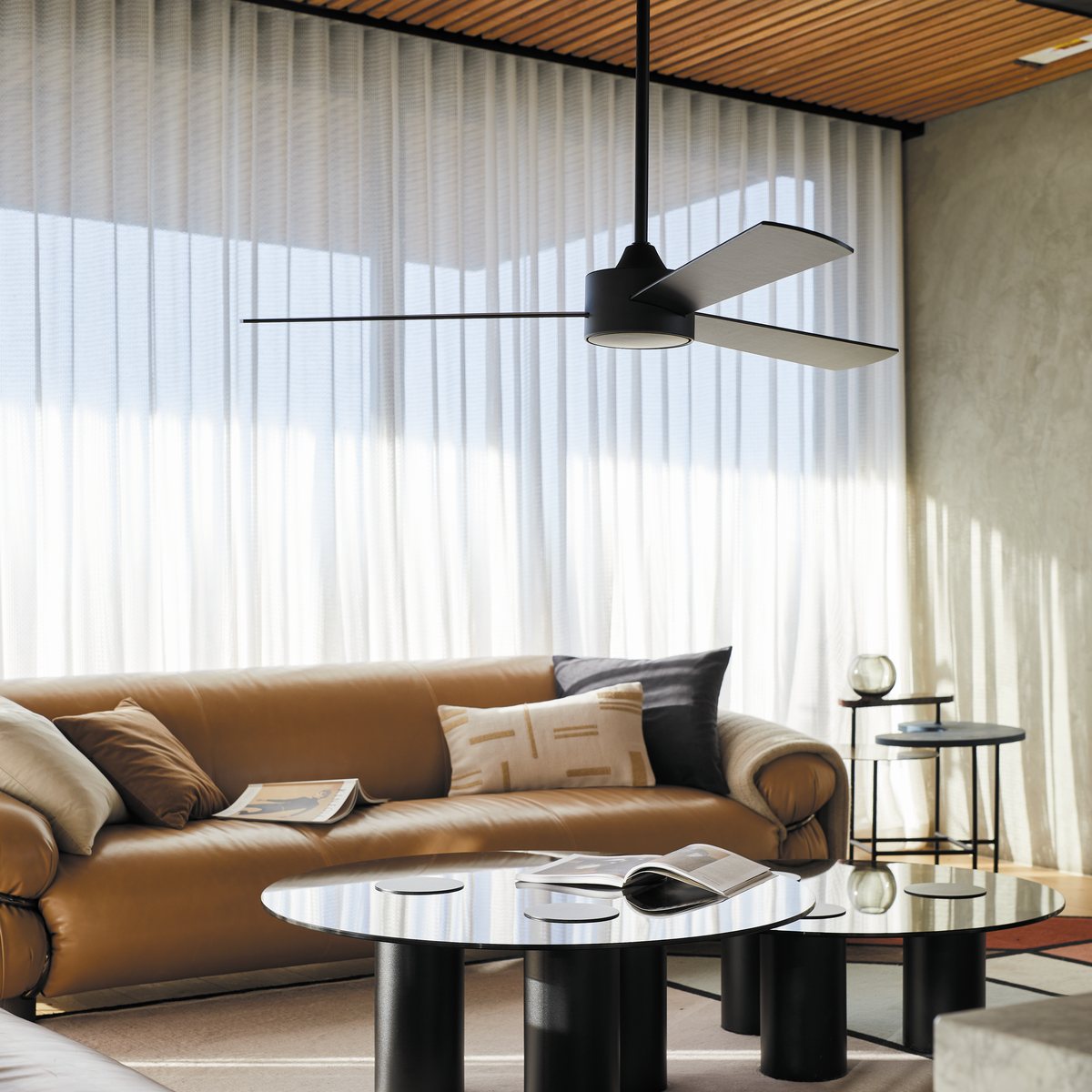 Black three-blade ceiling fan above a tan leather sofa and round black coffee tables in a living room with sheer curtains and a timber slat ceiling
