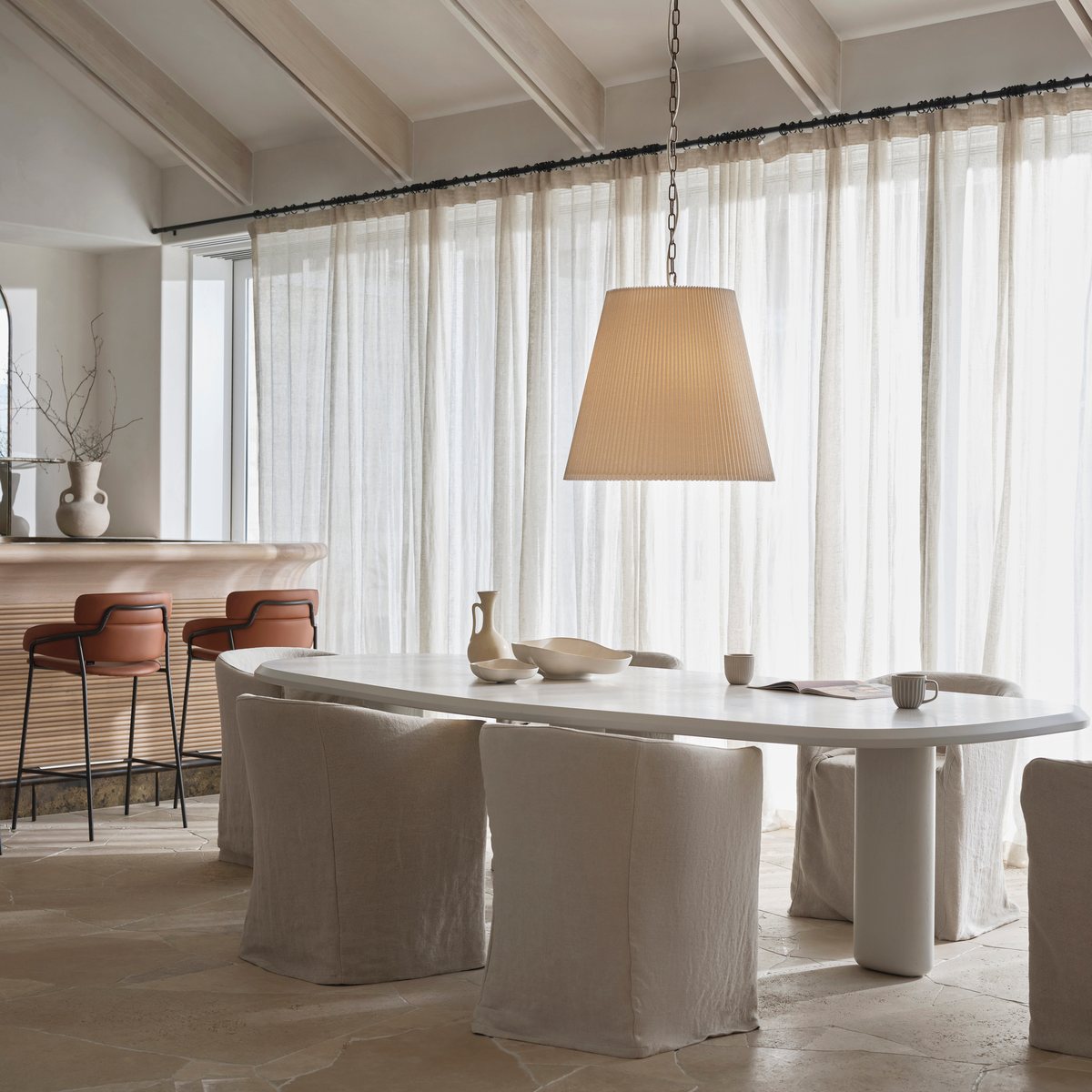 Pleated linen pendant light hanging above an oval white dining table with linen-covered chairs in front of sheer curtains