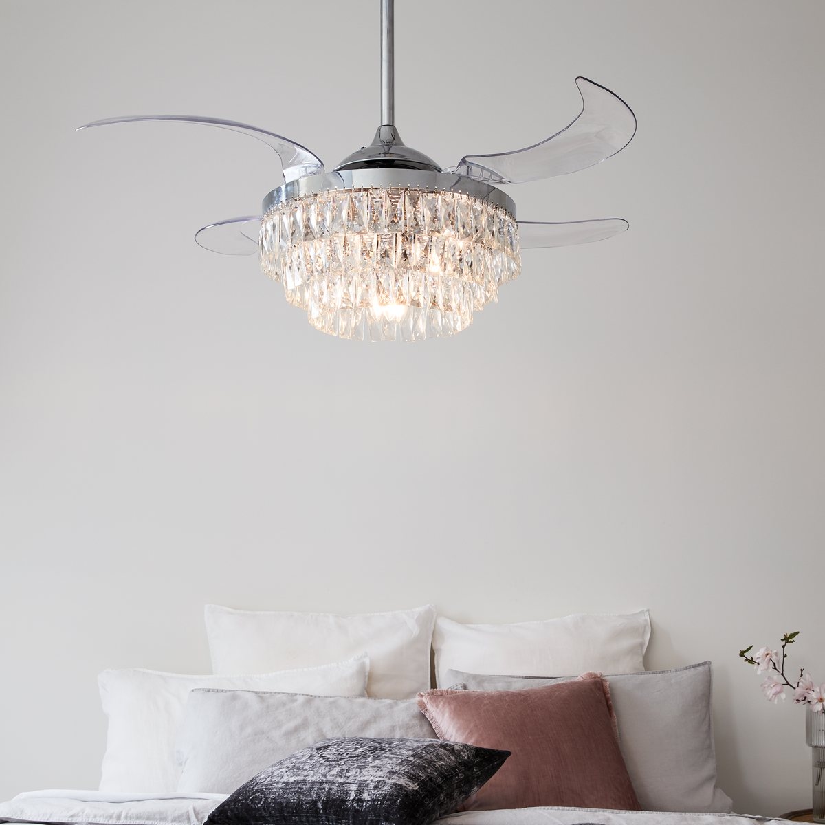 Chrome ceiling fan with clear retractable blades and a crystal light fitting above a bed with white and blush pillows