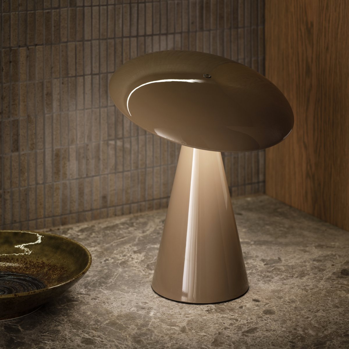 Taupe table lamp with a wide domed shade on a stone surface in front of a brown tile wall