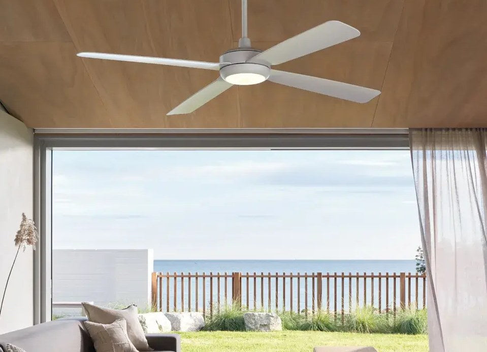 White four-blade ceiling fan with a light under a timber ceiling, above a living room that opens to a lawn and the sea