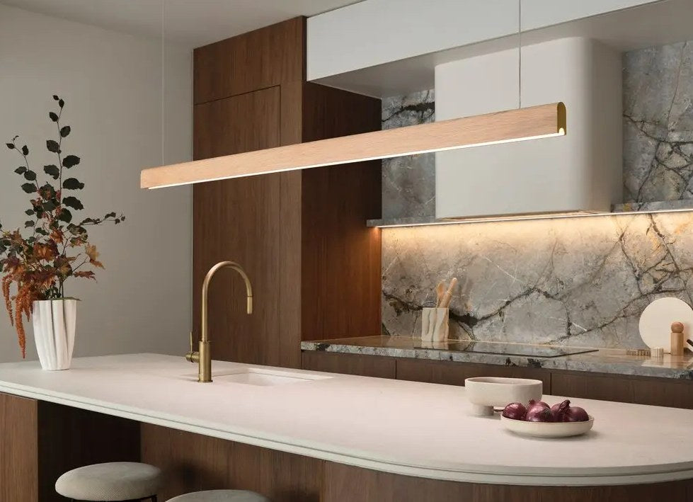 Long timber linear pendant light above a curved marble kitchen bench with a brass tap and bar stools