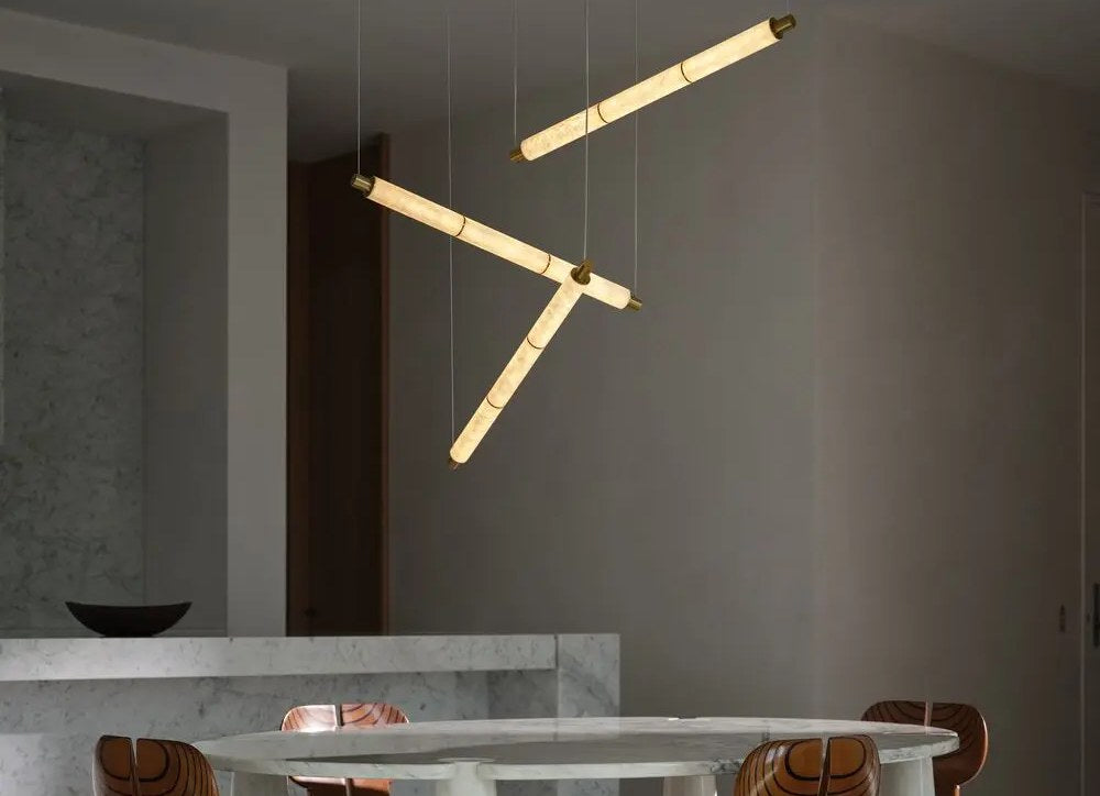 Three slim glowing linear pendant lights hung at angles above a marble dining table with sculptural wooden chairs